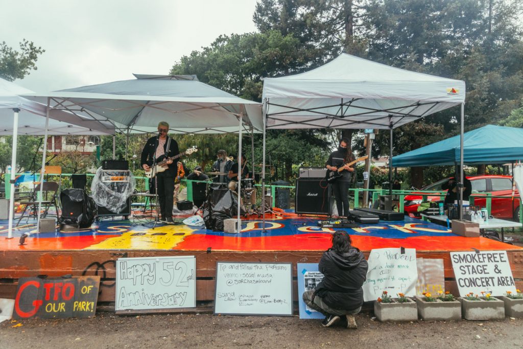 Berkeley-based pop punk band, Sarchasm, plays on the red and blue People's Park stage. The band plays beneath easy ups on the stage to protect themselves and their instruments from the rain.