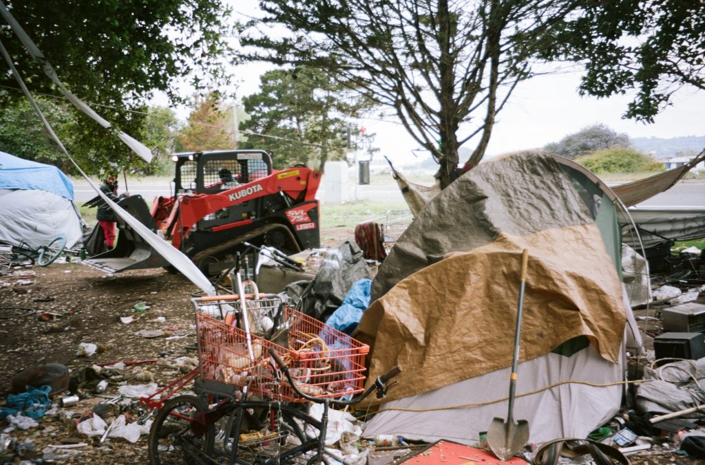 A shovel rests against a tent. Garbage surrounds the tent, and a volunteer can be seen cleaning up in the background.