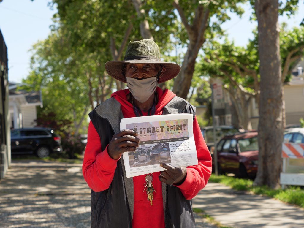 Kevin Aikens, a Black man wearing a red sweatshirt and a floppy hat, holds up a copy of Street Spirit.