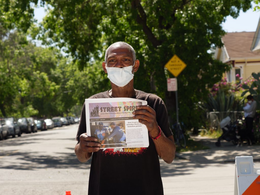 Bobby, a Black man in a black shirt, holds up a copy of Street Spirit with a seemingly residential street behind him.