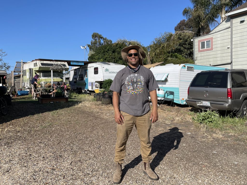 Adam Garrett-Clark stands in the middle of the Neighborship RV community. RVs and cars can be seen behind him. Hr wears work pants and boots, and stands squarely in front of the camera smiling.