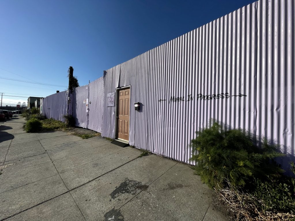 A powder purple, corrugated fence has a brown wooden door in the middle. This is the entrance to the Neighborship community. The words "Mural in progress" are painted on the fence.
