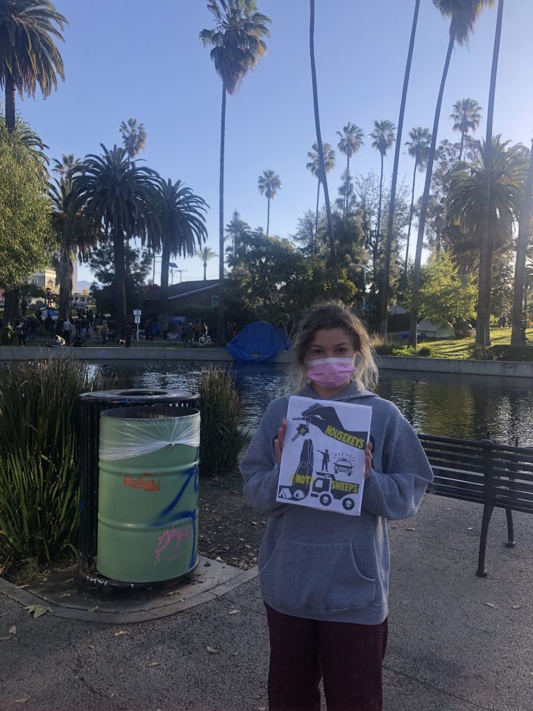 A masked protestor stands in a Los Angeles public park, with palm trees and a lake in the background. She holds the "housekeys not sweeps" poster in her hands.