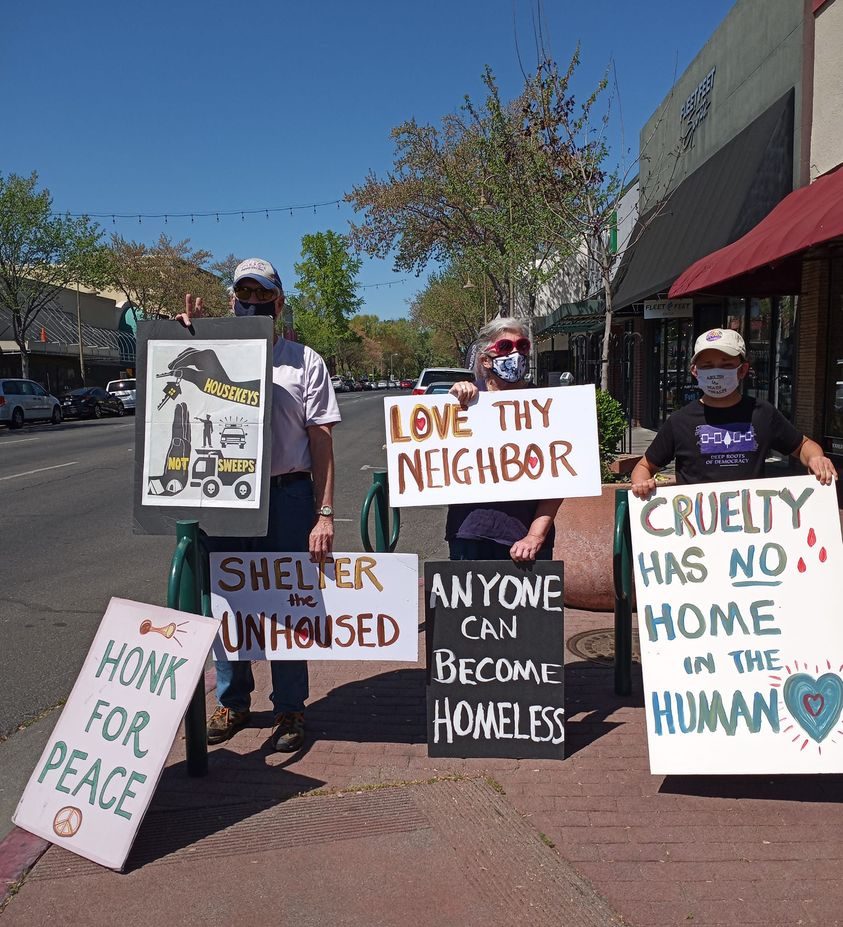 A group of masked protestors stand on the sidewalk in Chico, CA, with signs that say "love thy neighbor," "Shelter the unhoused," "Honk for peace," and "housekeys not sweeps."