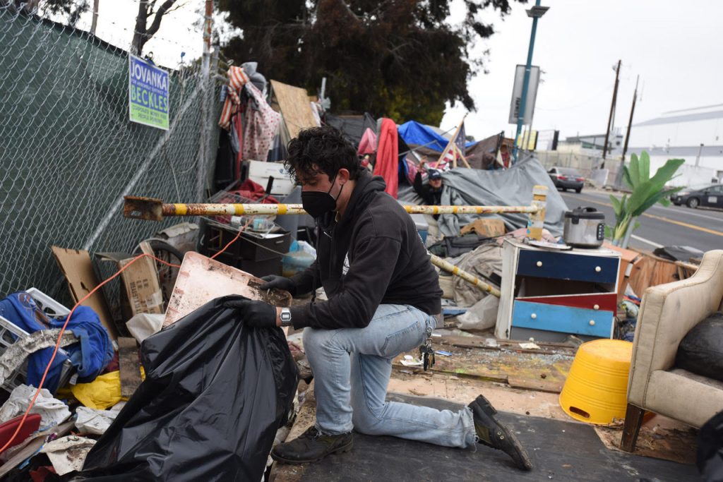 Ian Cordova Morales kneels amidst scattered trash and encampment resident possessions, loading things into black trash bags.