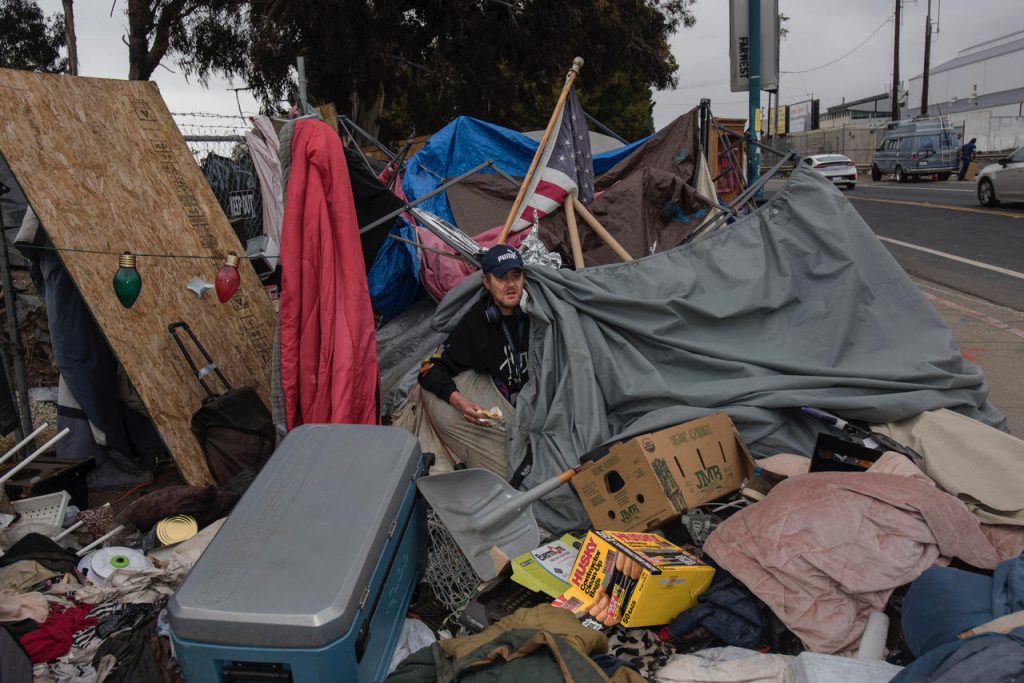 Gabe pokes his head out of his tent. He is surrounded by a big pile of his belongings, as well as trash bags for moving. An American flag pokes out of the refuse behind him. Red and green string lights are attached to his tent.