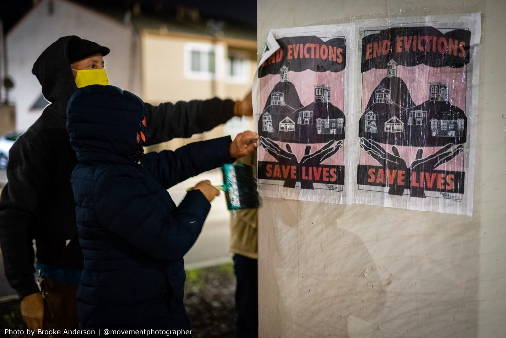 Two hooded people use wheatpaste to paint a poster that reads "end evictions, save lives" onto a concrete pillar so that it sticks.