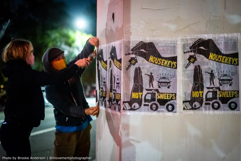 Two hooded people use wheatpaste to paint a poster that reads "housekeys not sweeps" onto a concrete pillar so that it sticks.