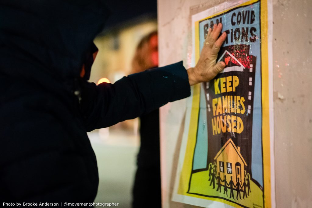A hooded figure pushes their hand flat against the "Ban covid evictions, keep families housed" holding it up against a concrete pillar, in preparation to use wheatpaste to stick the poster to the pillar. 