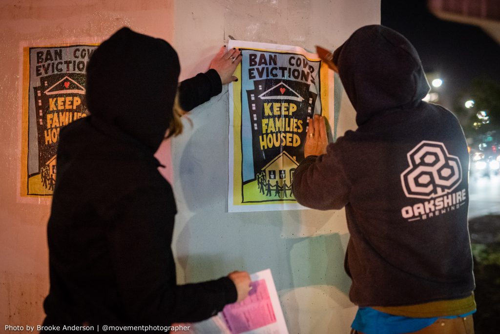 Two hooded people use wheatpaste to paint a poster that reads "ban COVID evictions, keep families housed" onto a concrete pillar so that it sticks.
