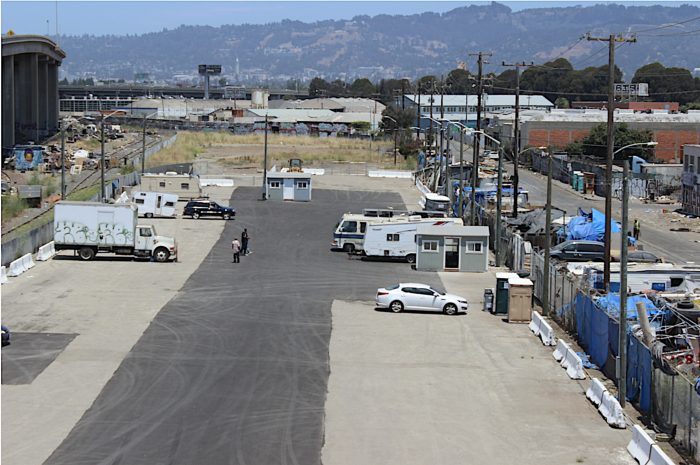 A nearly empty parking lot where the wood street safe parking program takes place. Six vehicles can be seen in the photo and an encampment presses up against the fence that separates the parking lot from the street.