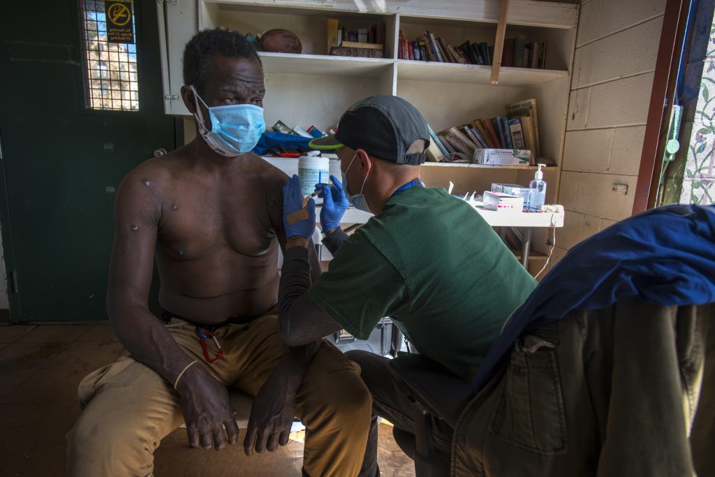 A shirtless man wearing a blue surgical mask gets the COVID vaccine. He has some scars on his chest.