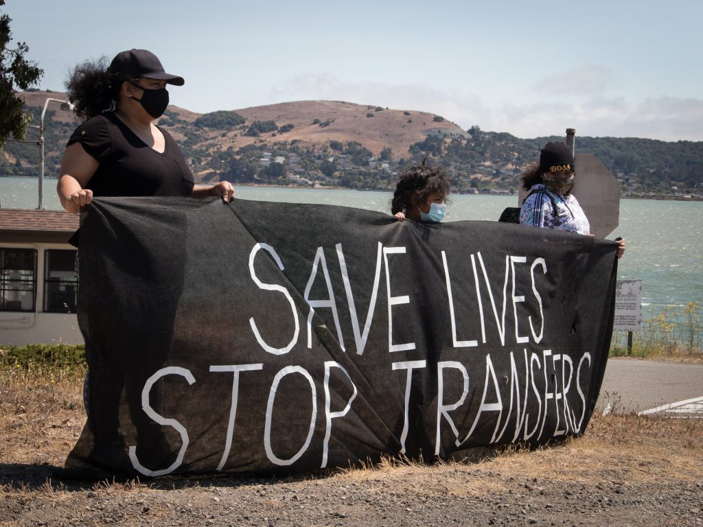 Three people hold a banner that says "save lives stop transfers" with San Quentin and the ocean in the background.