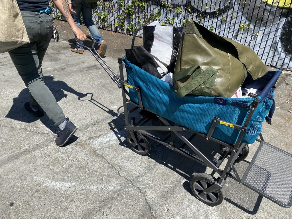 A person wearing grey jeans and black sneakers pulls a blue cart containing bags of Narcan and other harm reduction supplies.