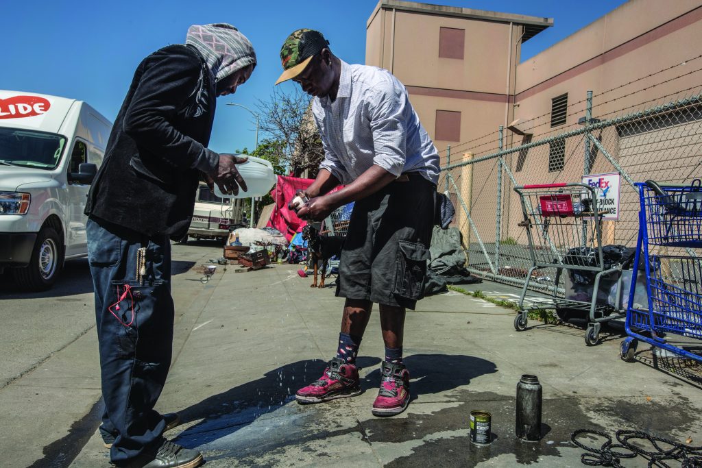 One man washes his hands with a bar of soap while another pours water over his hands from a plastic gallon container. They are standing on the sidewalk and shopping carts and a dog can be seen in the background.