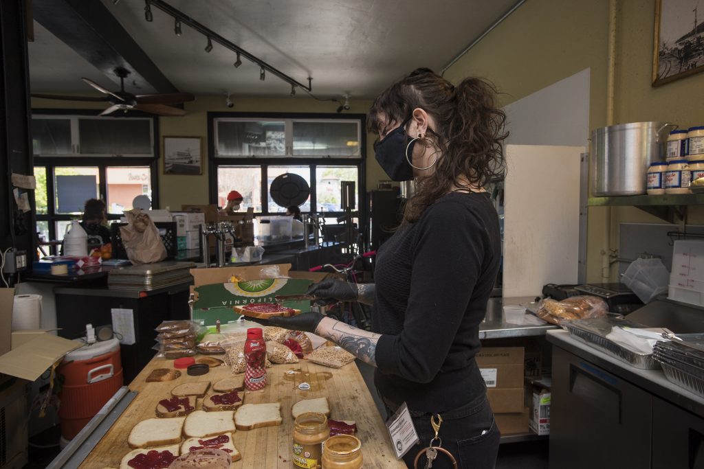 A woman in black spreads jelly on a piece of bread. Several pieces of bread with jelly and peanut butter lie on the table before her, waiting to be assembled into PB&J sandwiches.