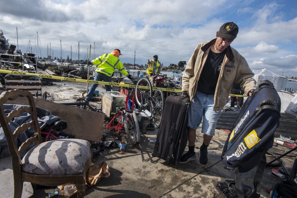 A man holding two suitcases walks away from his homeless encampment. Behind him, a pile of bicycles and old chairs sit behind a line of yellow caution tape. Two public works employees in neon vests work on removing items from the area.