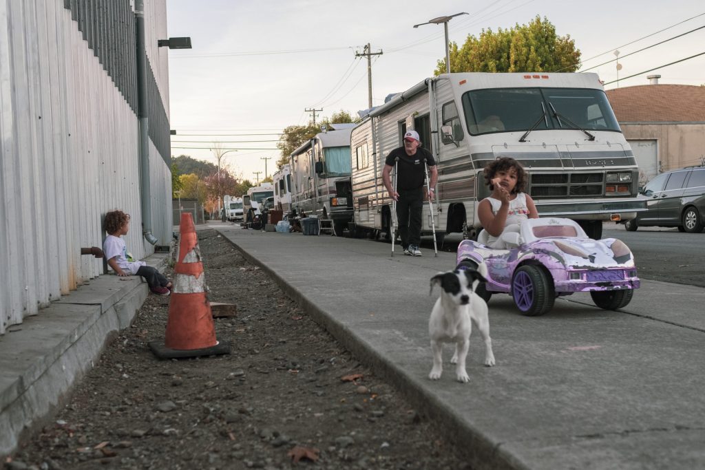 A little girl sits in a large toy car on the sidewalk outside of the RV where she lives. There is a puppy in the foreground in front of her, and behind her, another child sits on the ground leaning up against a building.  person with crutches stands in the background watching the children.