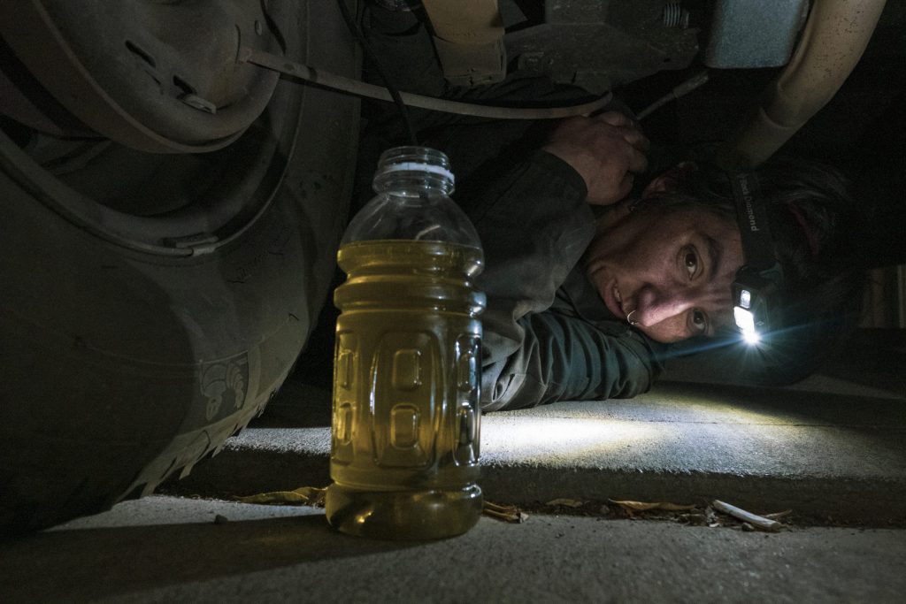 Amber Whitson shimmies herself beneath a vehicle to drain the brake fluid. In the foreground, an empty bottle is full of yellow brake fluid.