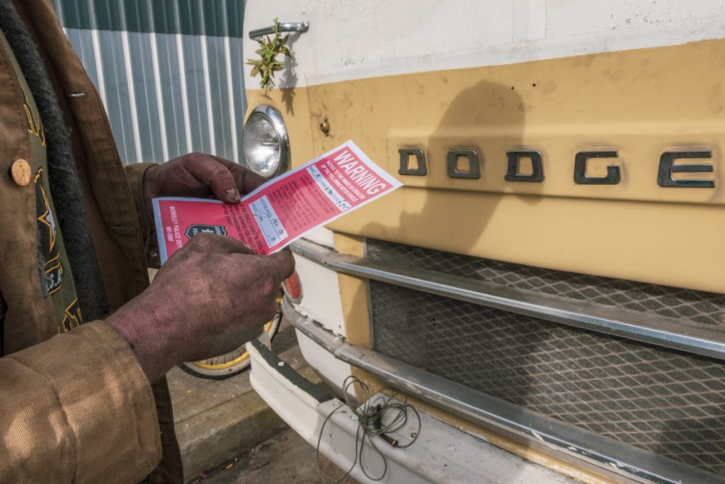 A person holding a red 72-hour tow notice. Their hands look dirty as they hold the notice, and their yellow dodge van can be seen in front of them.