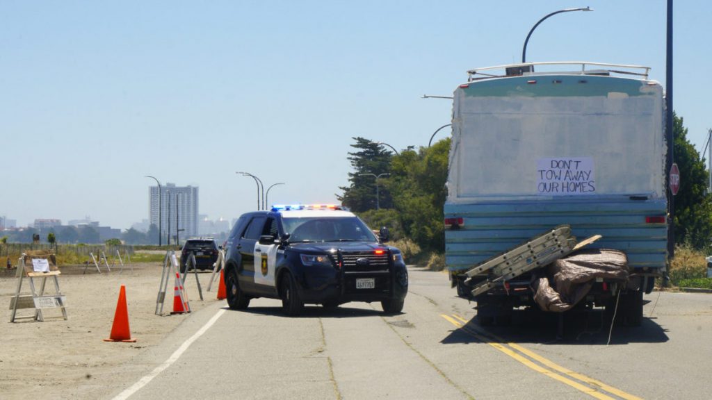 A police car blocks off a street near the Berkeley Marina. It is parked in the middle of the road next to an RV with a sign on the back that reads "don't tow away our homes."