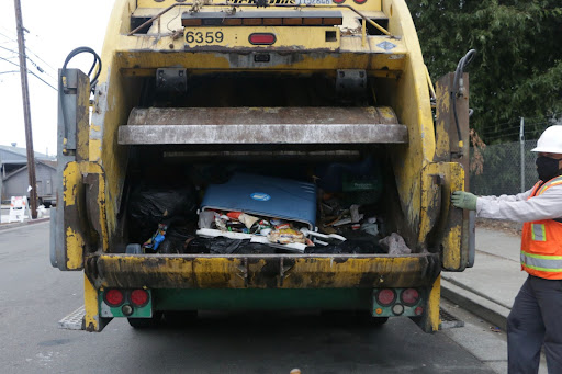 An open garbage truck with a full cooler piled amongst the trash. A public works employee in an orange neon vest can be seen in the foreground.