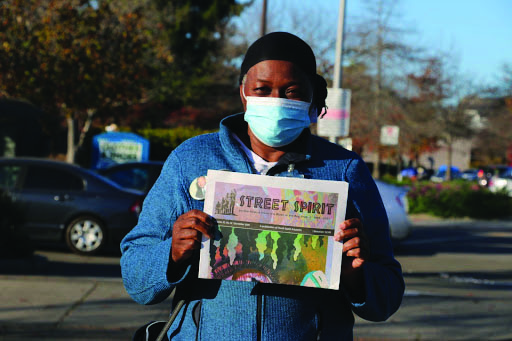 Vinola Stallings, a middle aged Black woman wearing a head wrap and a surgical face mask, holds up a copy of Street Spirit.