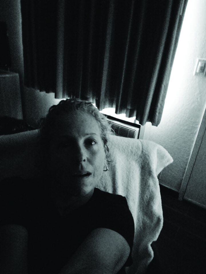 A black and white selfie of Laura, a white woman who looks to be in her 40s, sitting in a motel room.