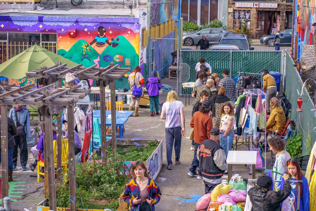 A group of people selling colorful wares set up in a lot behind Youth Spirit artworks, surrounded by murals.