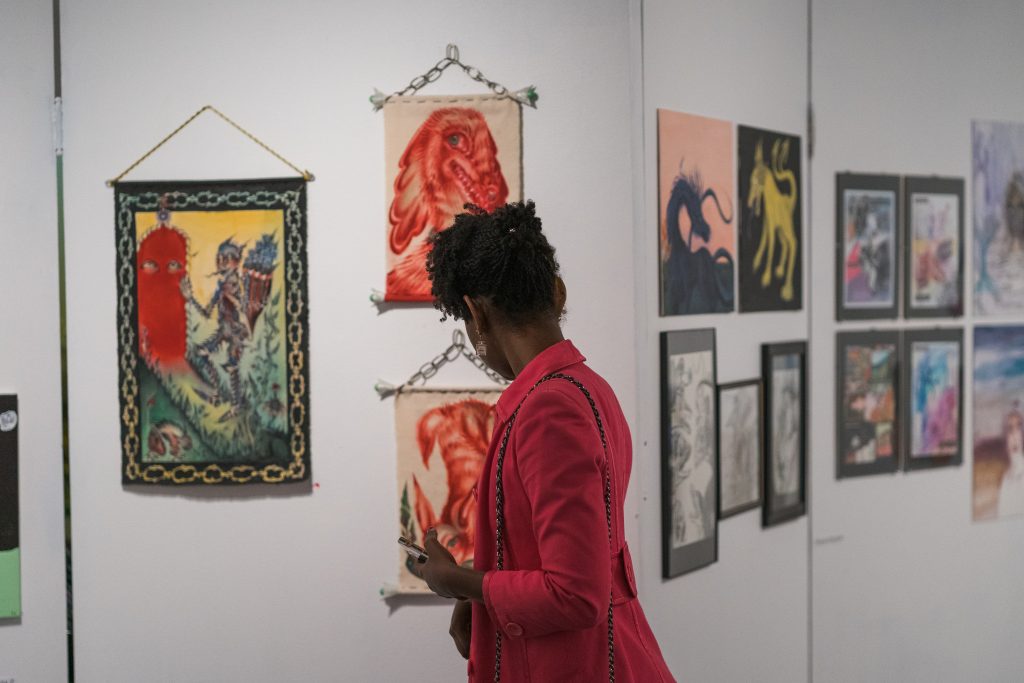 A Black woman in a red coat looks at artwork on the walls in the gallery with her back facing the camera.