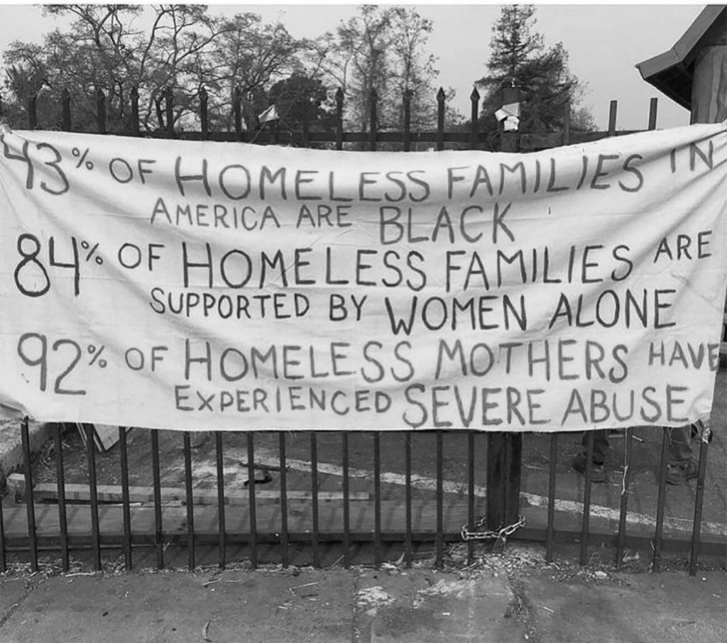 A protest sign hanging on a fence reads: "43 of homeless families in America Are Black. 84 of homeless families are supported by women alone. "92 of homeless mothers have experienced severe abuse"
