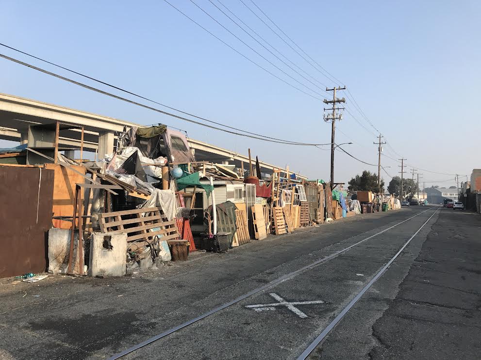 A photo of an encampment that stretches down a long block, with lots of self-built structures facing the street. The structures are made out of wood and pallets.
