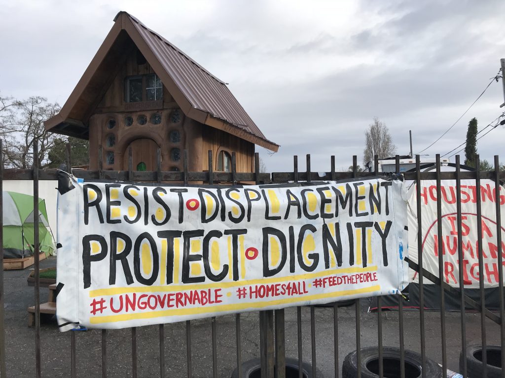 A banner hangs on the fence for Housing and Dignity Village that reads "resist displacement protect dignity #ungovernable #homes4all #feedthepeople." In the background, a wooden tiny home and a tent can be seen. The tent bears writing that says "housing is a human right." 