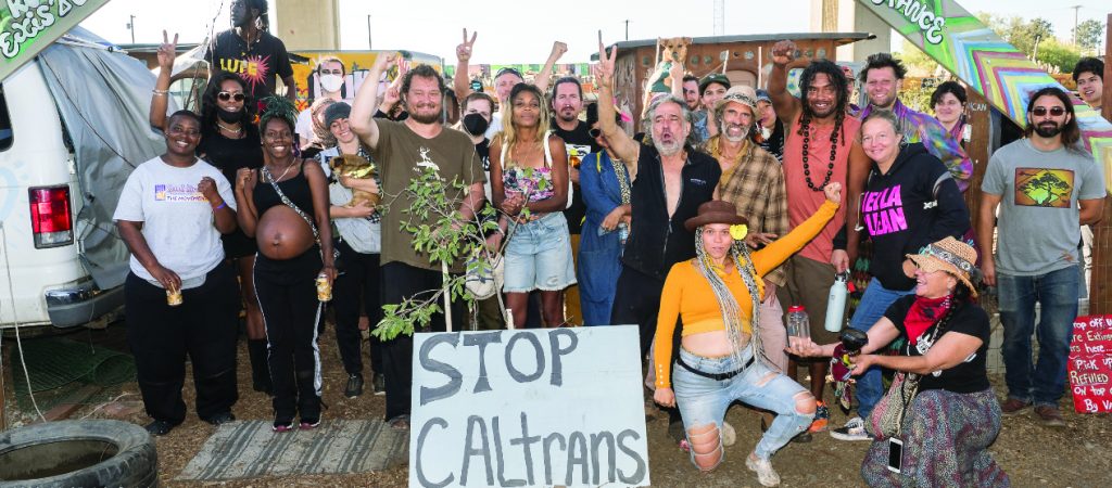 A large group of people gather together and smile, or hold peace signs up. In front of them a protest sign reads "stop Caltrans."