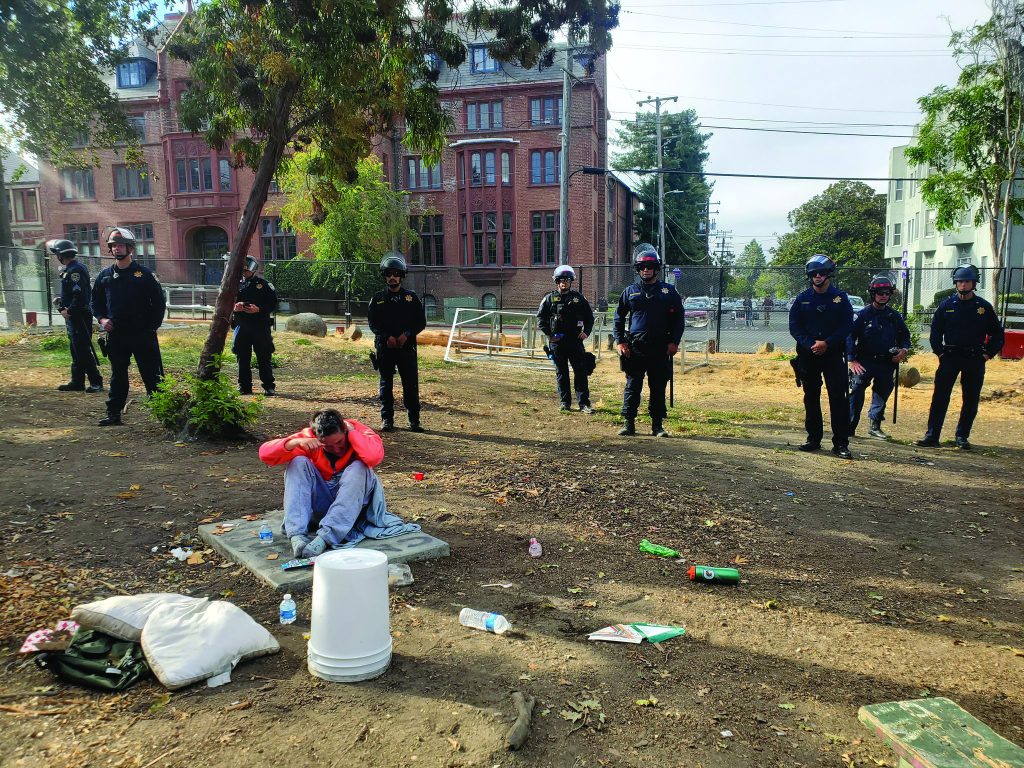 An unhoused woman sits on the ground with her head leaning over her knees and her hands on her ears. Nine police officers stand in a line behind her.