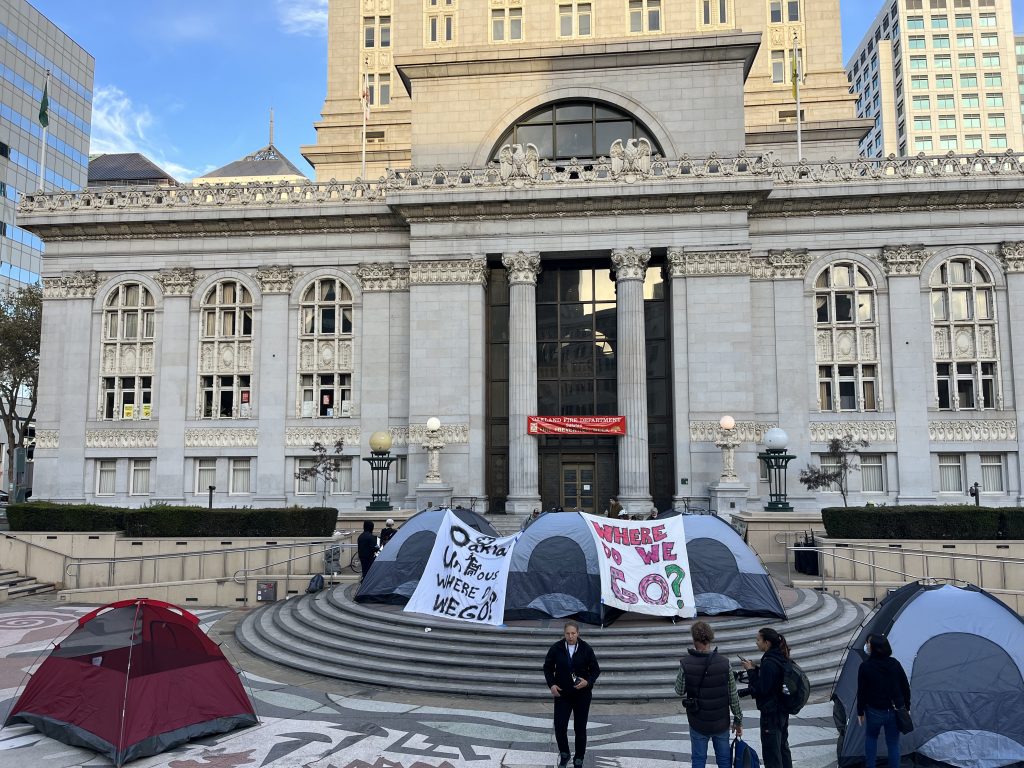 Five tents are set up around the front steps of Oakland City Hall. Two banners hang from two of the tents. One of them says "Where do we go?" and the other says "Oakland unhoused, where do we go?"