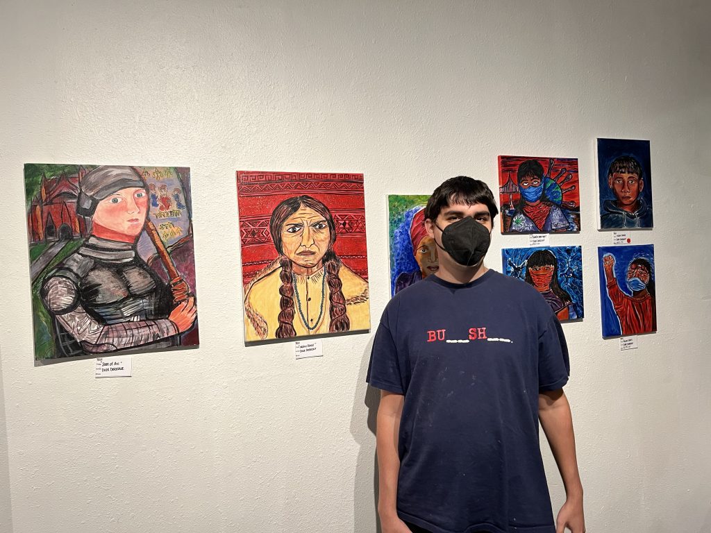 Delacour—a white man with dark brown hair—stands in front of a wall of portraits that feature vivid colors, especially reds and blues.