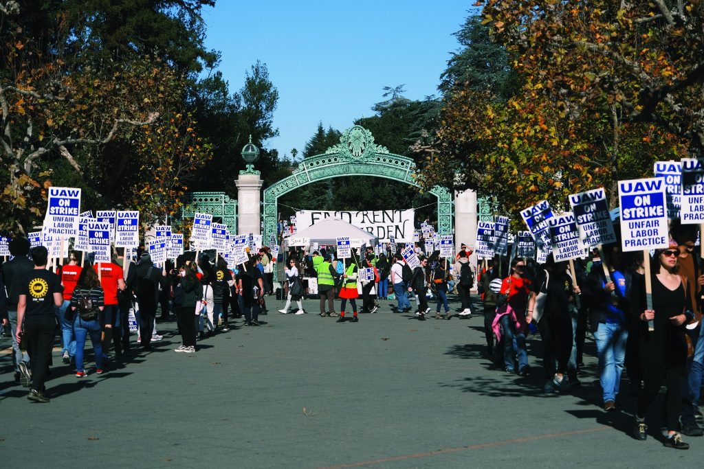 I swarm of striking students underneath and around UC Berkeley's Sather Gate with signs that say "UAW on strike, unfair labor practice." In the background a large banner reads "end rent burden"