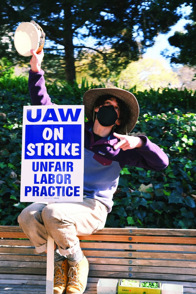 A striking student wearing a sunhat and holding a UAW protest sign and plays the tambourine. 