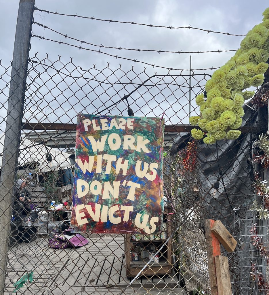 A photograph of a chain link fence with a multi-colored poster on it. In front of a multi-colored, rainbow-ish backdrop are the words "please work with us don't evict us". 