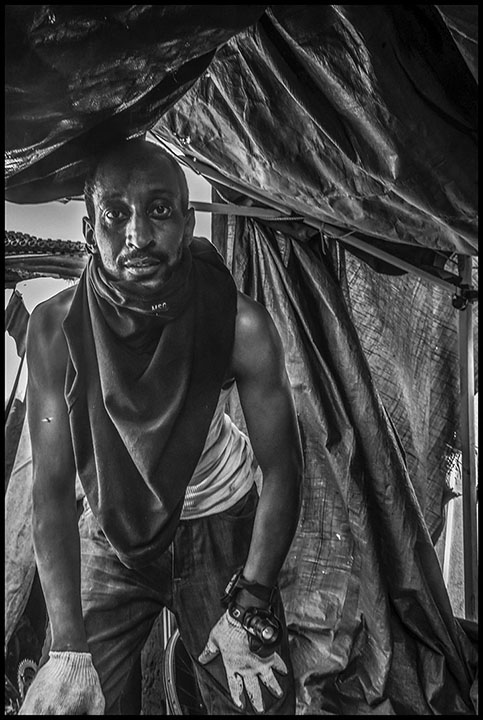 A black and white photo of a Black encampment resident within his dwelling, with a what looks like a tarp hanging above him as the roof.