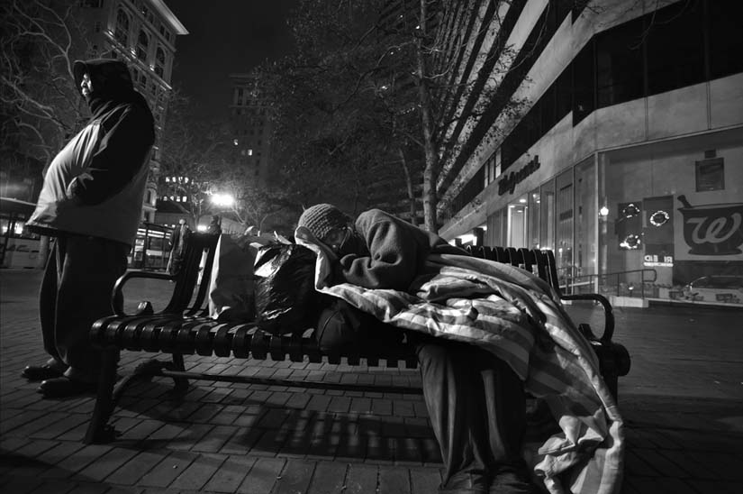 In Oakland, a homeless woman sleeps on a bus bench while a homeless man watches protectively over her. Other homeless people brought her food and blankets. David Bacon photo