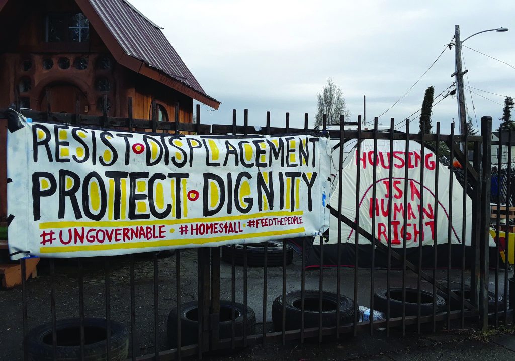 A banner hangs from a fence. It reads "Resist displacement protect dignity #ungovernable #homes4all #feedthepeople"