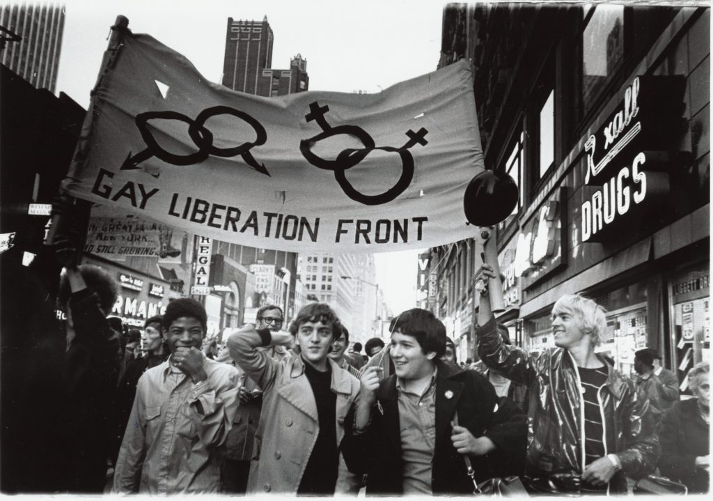 Gay Liberation Front marches on Times Square, New York, 1970. A black and white photo of a group of people in a crowd. They hold up a big banner that reads "gay liberation front."