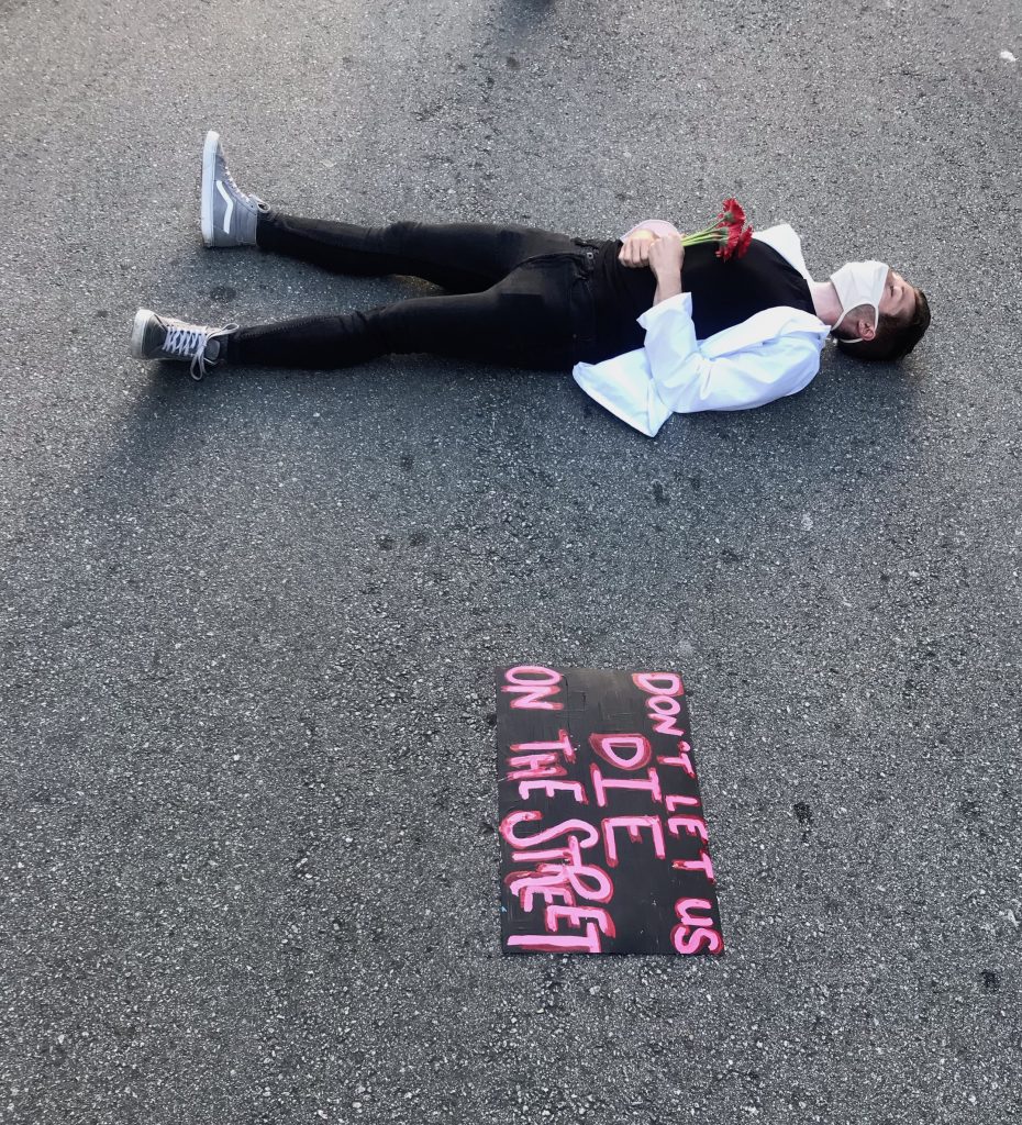 A protestor in a white coat lies on the ground with flowers in their hand, with a sign to their left that reads "don't let us die on the street"