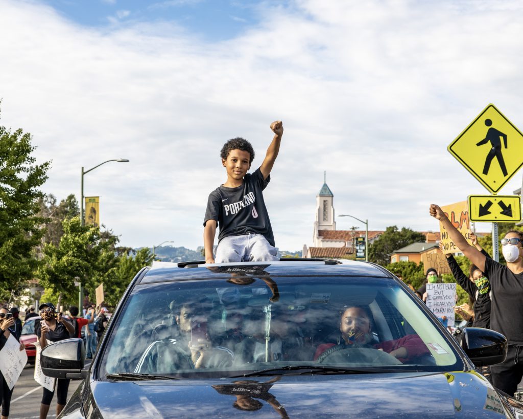 A young black boy sits on top of a car, through the sun roof, holding up the fist of black power. Around him in the street protestors cheer and raise their fists in return.