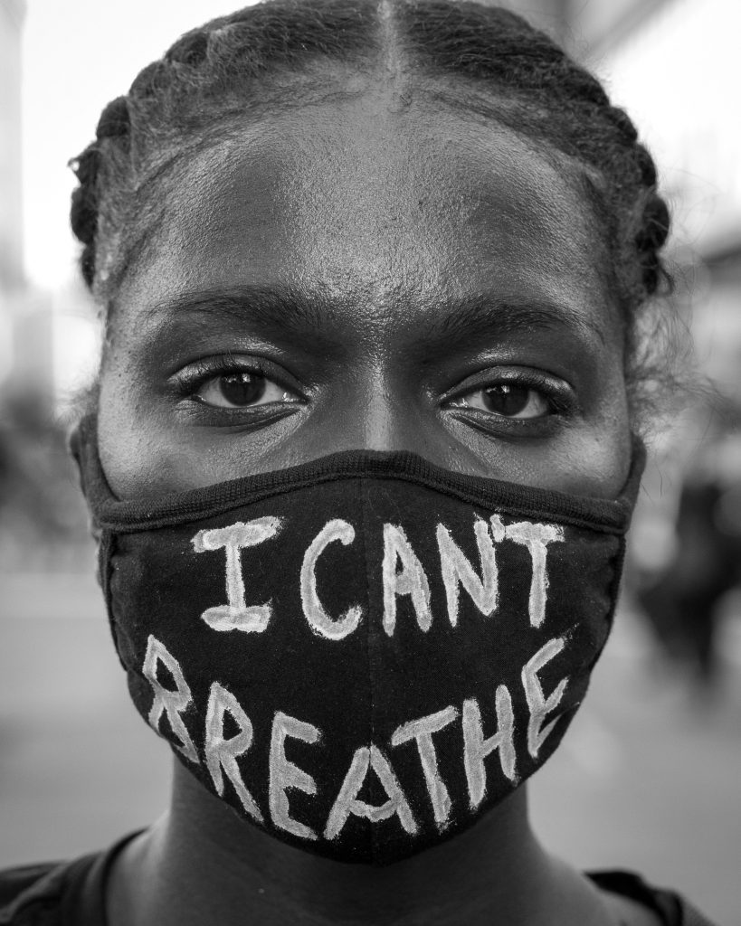 A close up, black and white portrait of a black woman with braids in her hair. Only her eyes can be seen, because she is wearing a dark face mask. On the mask she wrote the words, "I can't breathe."