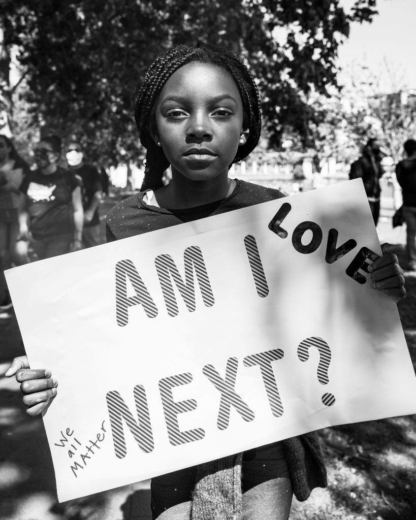 A young Black girl holds up a sign that says "Am I next?" and "love." he image is in black and white. The image is in black and white.  