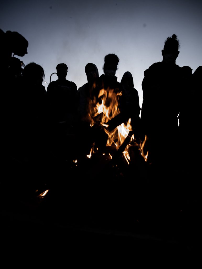 Another night time shot. Seven or so people gather around a fire, which burns bright at the center of the shot. The fire is orange, but you can only see silhouettes of the people, who are all wearing different hair styles and textures. Behind them the sky is navy blue as though the sun has just set.
