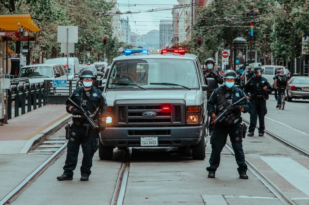 On San Francisco's Market Street, two police officers stand guard in front of a police van with its lights on. They are armed with rubber bullet weapons.